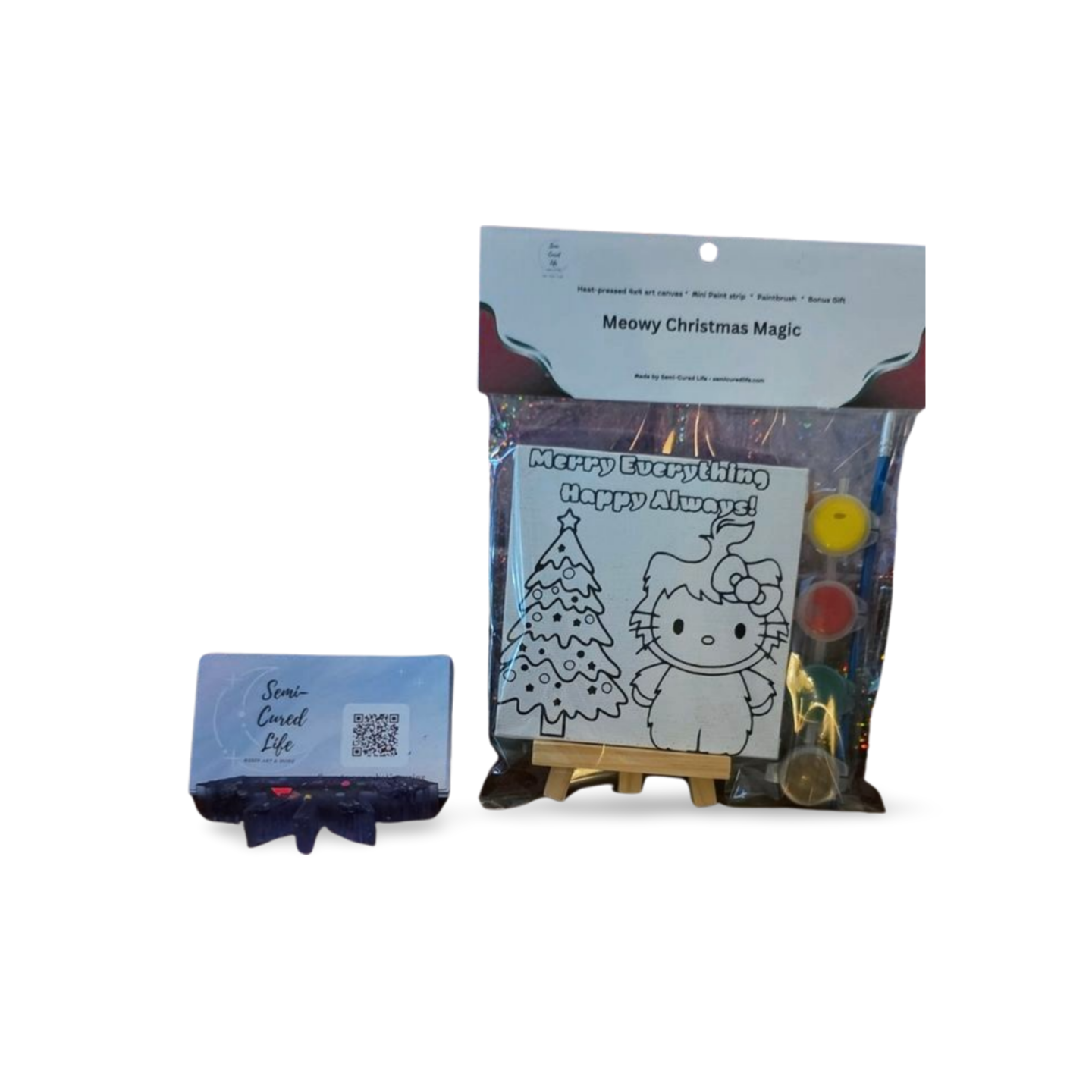Holiday Kitty mini canvas paint kit in clear bag with topper, showing Christmas tree and kitty design, paint pots, brush, and bonus gift on a small easel.