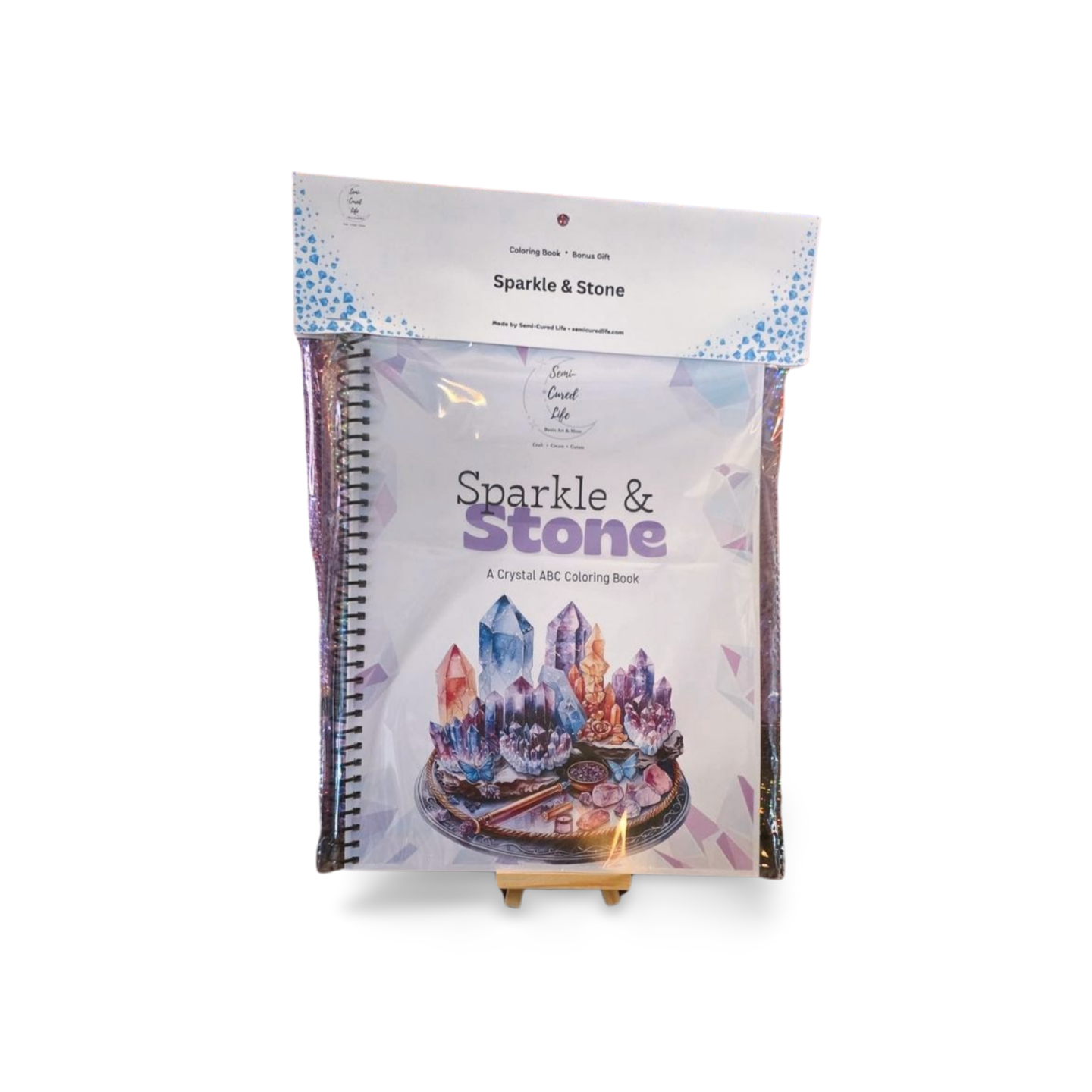 Sparkle & Stone crystal ABC coloring book packaged in a clear bag with branded topper, showing the spiral-bound book cover with pastel crystal cluster artwork on a tray.